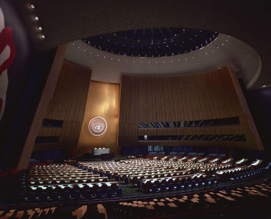 The United Nations General Assembly hall, empty, with the UN emblem above the rostrum.